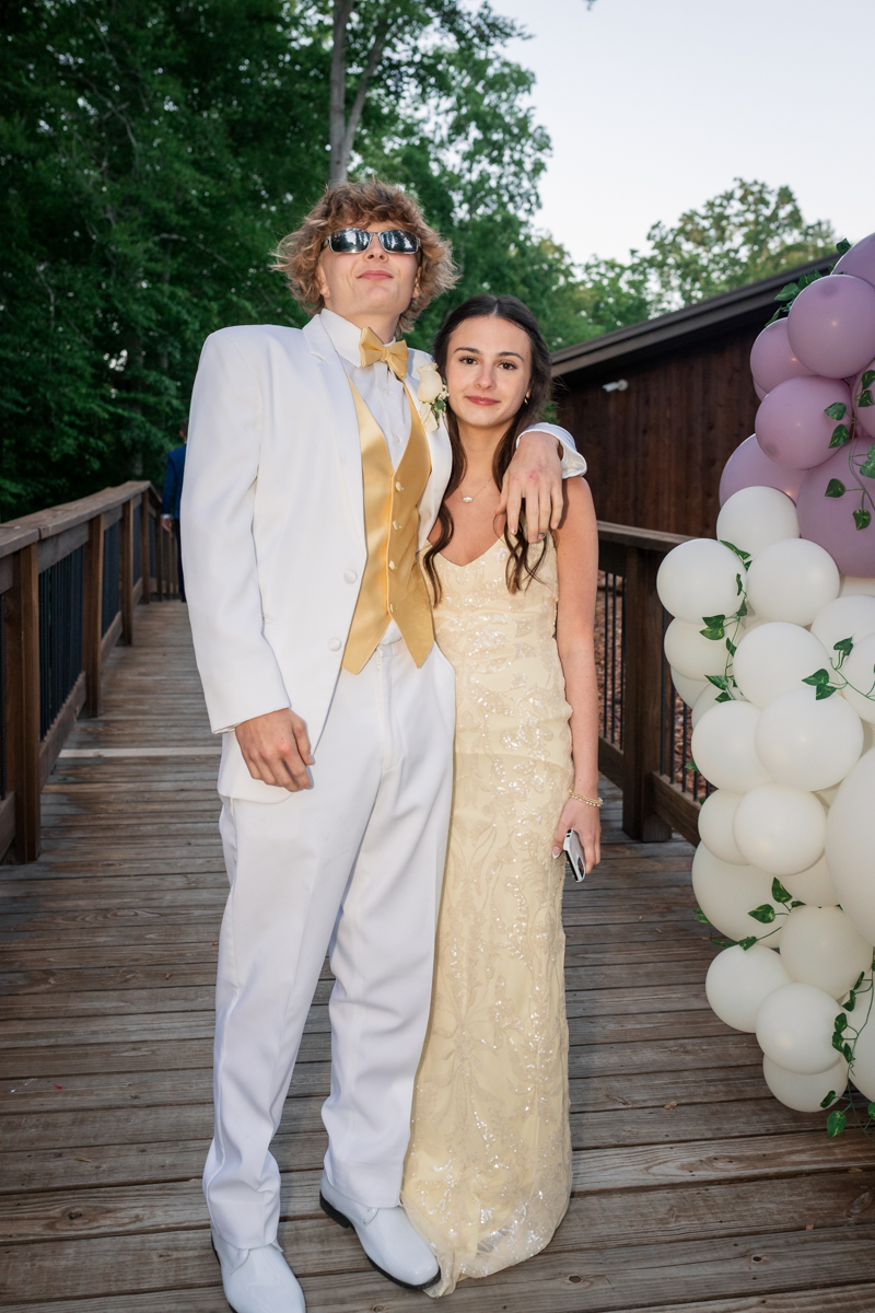 Couple posing on a wooden walkway at a formal event; man in a white suit with gold vest and sunglasses, woman in a champagne gown with arm around his shoulder.
