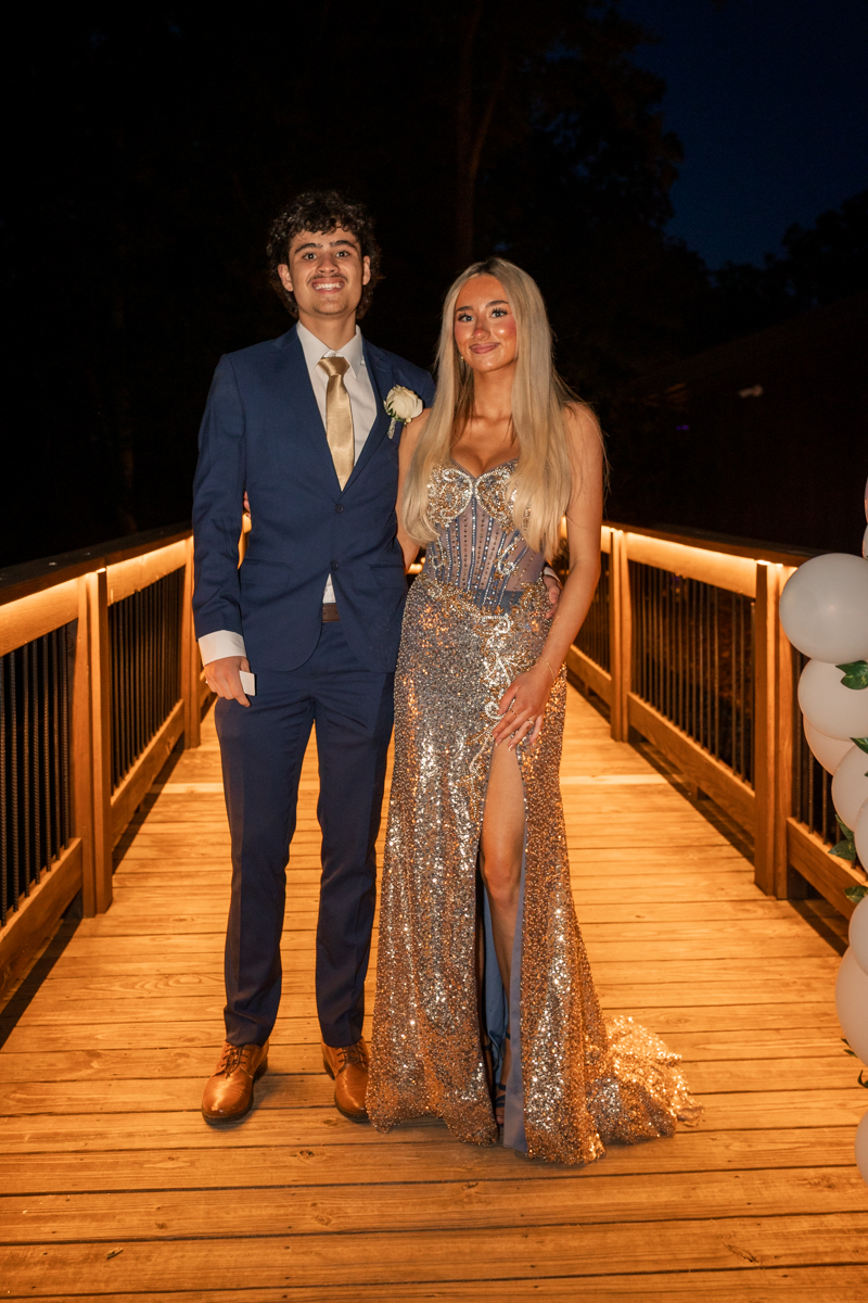 Couple in formal attire posing on a lit wooden walkway at night, man in blue suit and tan tie, woman in a sparkling gown with a high slit.