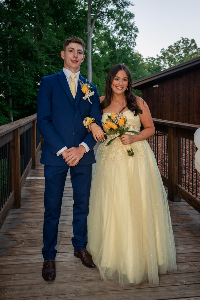 Young couple in formal wear pose on a wooden deck; the man wears a blue suit with a yellow boutonniere and the woman a pale yellow gown holding a bouquet of yellow roses.