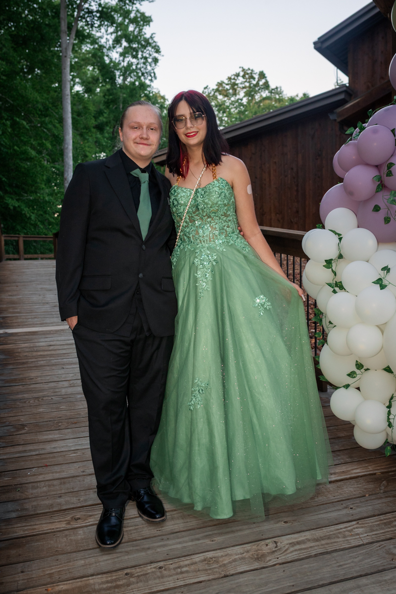 Two people in formal attire posing on a wooden deck: a man in a black suit and green tie next to a woman in a mint green gown.