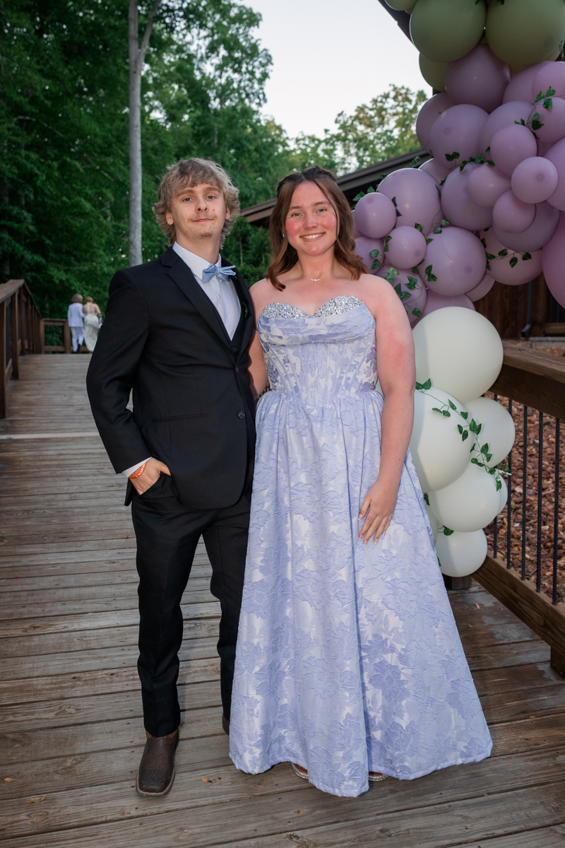 Young man in a black suit and blue bow tie poses with a smiling woman in a lavender strapless gown on a wooden deck beside a pastel balloon arch outdoors.