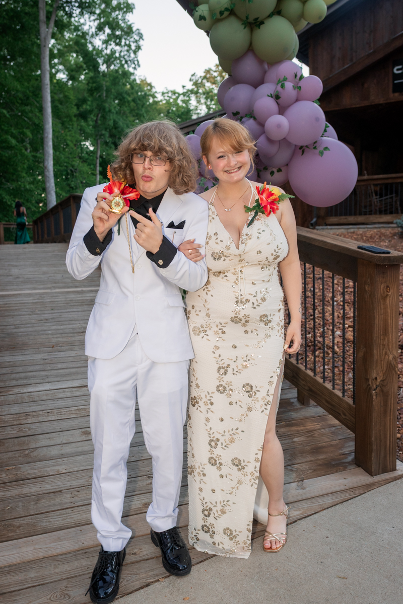 Young couple in formal attire pose on a wooden deck; the man in a white suit displays a medal while the woman in a gold floral dress smiles, with pastel balloons in the background.