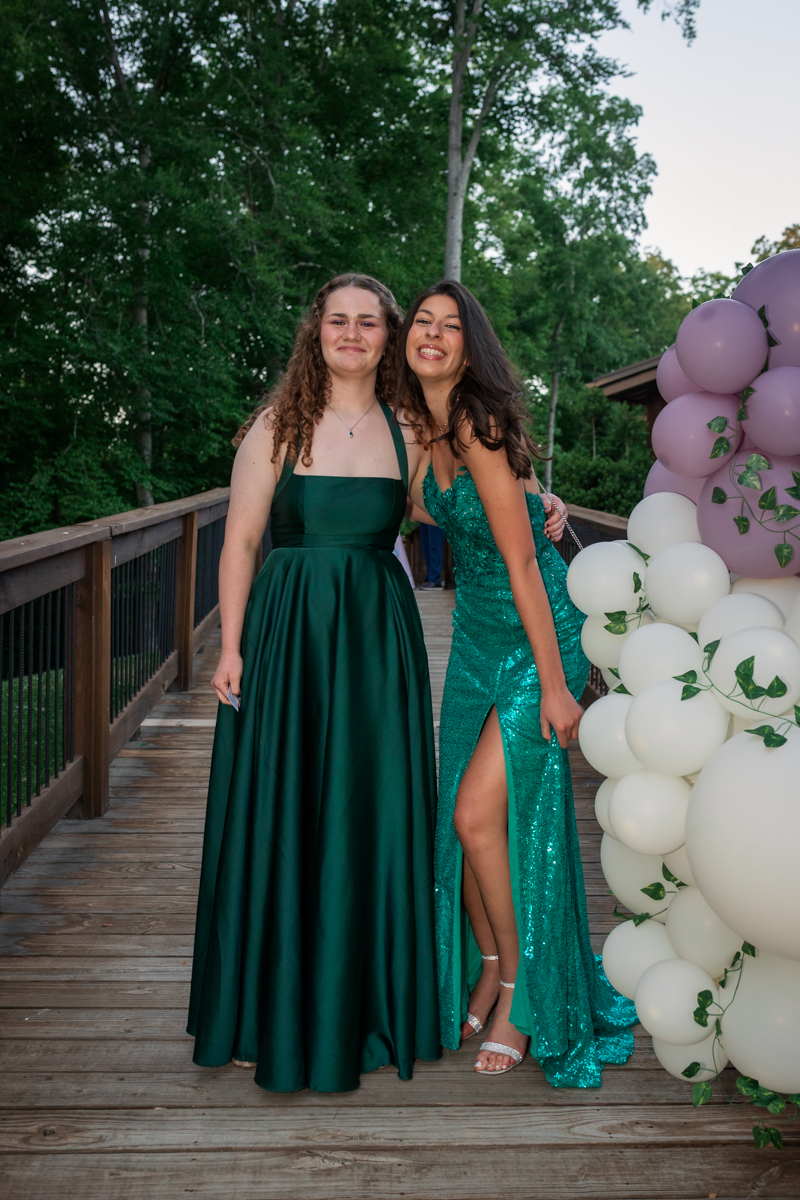 Two young women in formal gowns pose on a wooden deck next to a balloon arch, smiling at the camera.