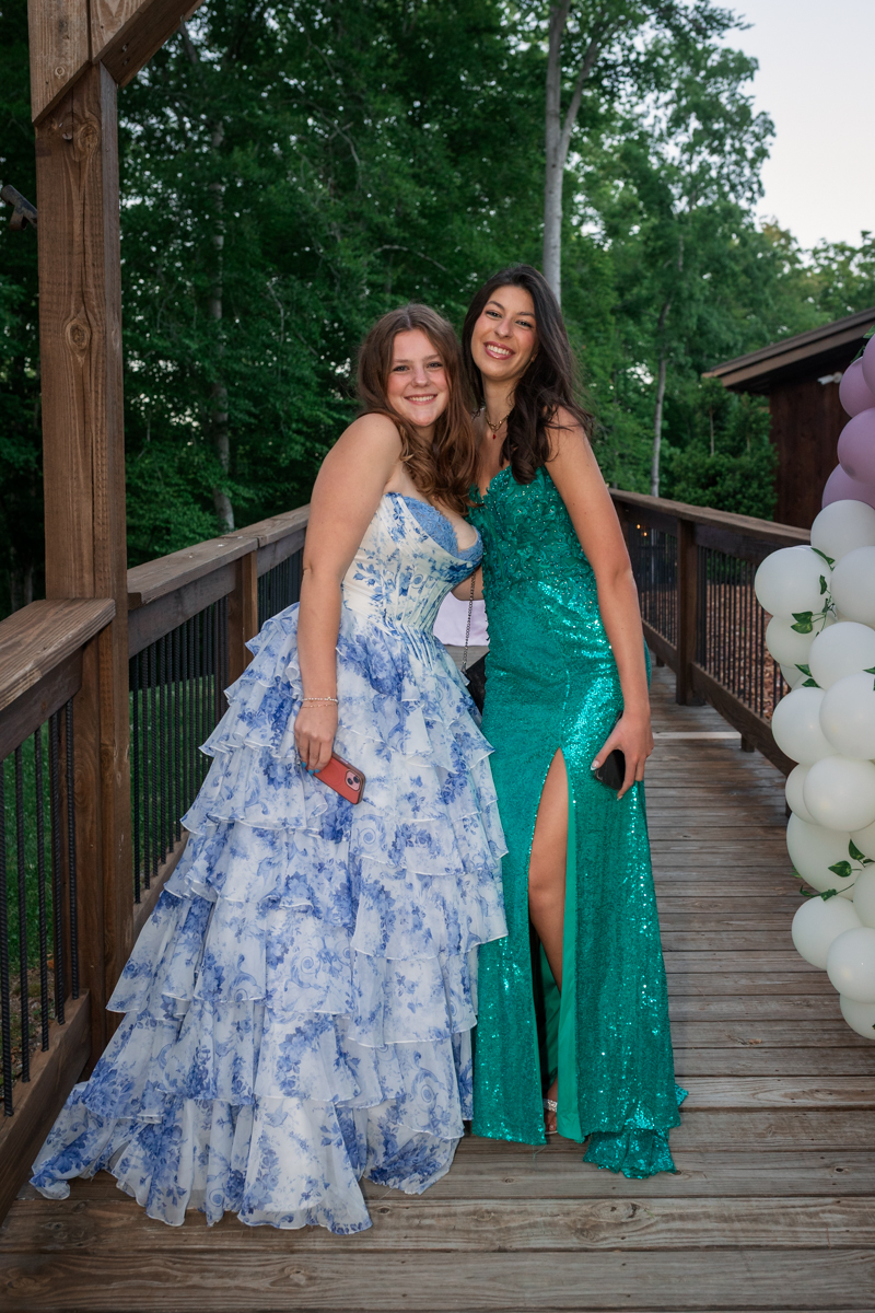 Two young women in formal gowns pose together on a wooden deck with balloons nearby, smiling at the camera.