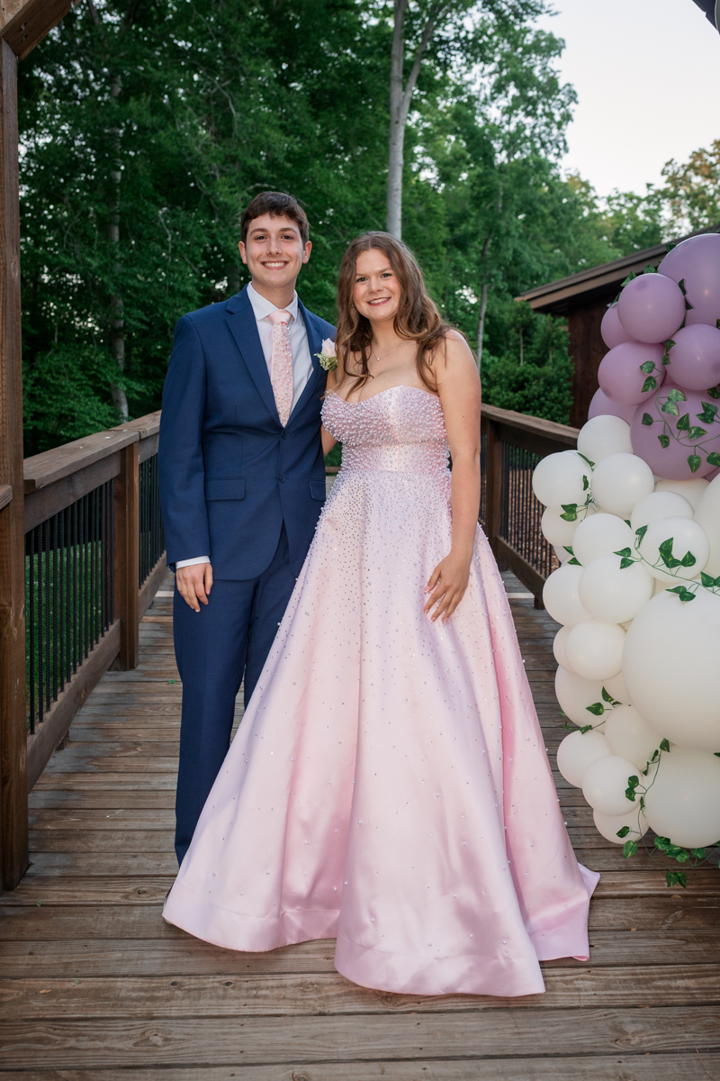 Smiling couple posing on a wooden deck, the man in a navy suit and pink tie, the woman in a light pink glittery gown with balloons nearby.