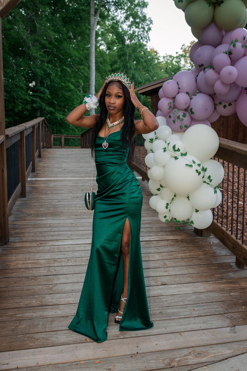Young woman in an emerald satin gown and crown posing on a wooden walkway with a pastel balloon arch beside her.