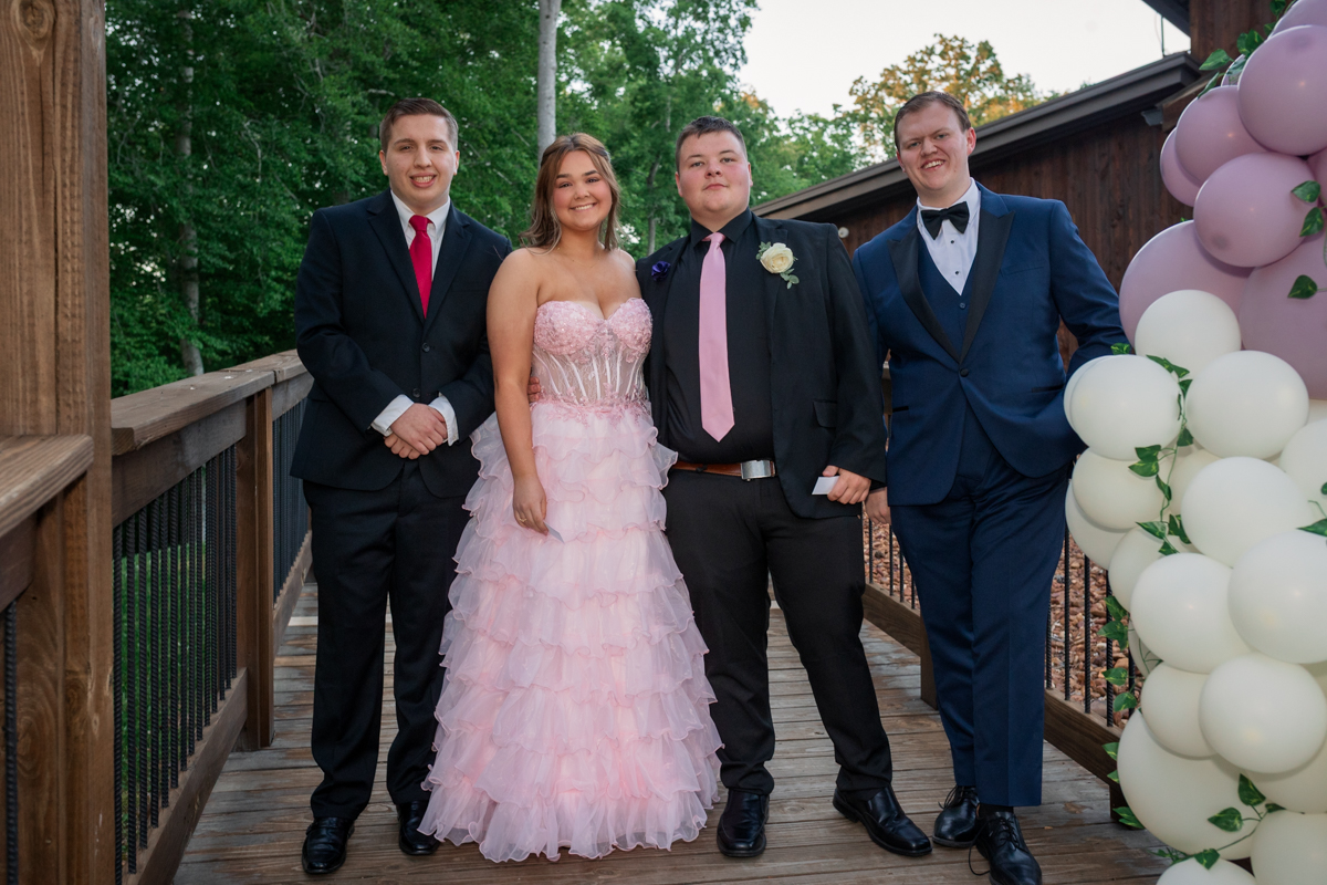 Group of four teens in formal wear posing on a wooden deck outdoors with a pink balloon arch nearby.