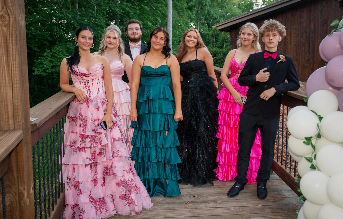 Seven teens in formal evening wear pose on a wooden deck outdoors, with greenery and pink/white balloons nearby.
