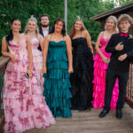 Seven teens in formal evening wear pose on a wooden deck outdoors, with greenery and pink/white balloons nearby.