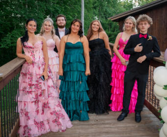 Seven teens in formal evening wear pose on a wooden deck outdoors, with greenery and pink/white balloons nearby.