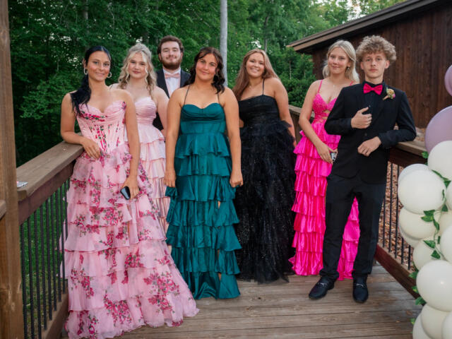 Seven teens in formal evening wear pose on a wooden deck outdoors, with greenery and pink/white balloons nearby.