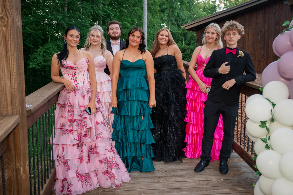Seven teens in formal evening wear pose on a wooden deck outdoors, with greenery and pink/white balloons nearby.