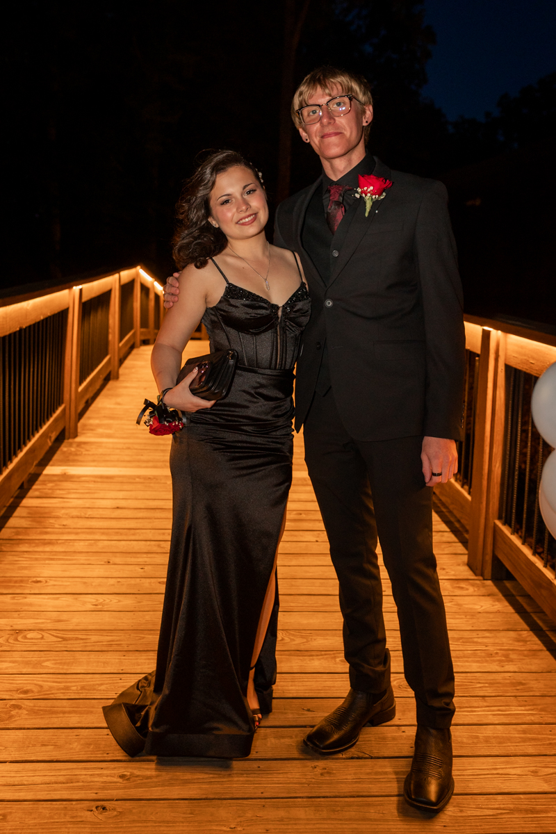 Couple dressed formally posing on a lit wooden bridge at night, woman in a black gown and man in a suit with a red boutonniere.