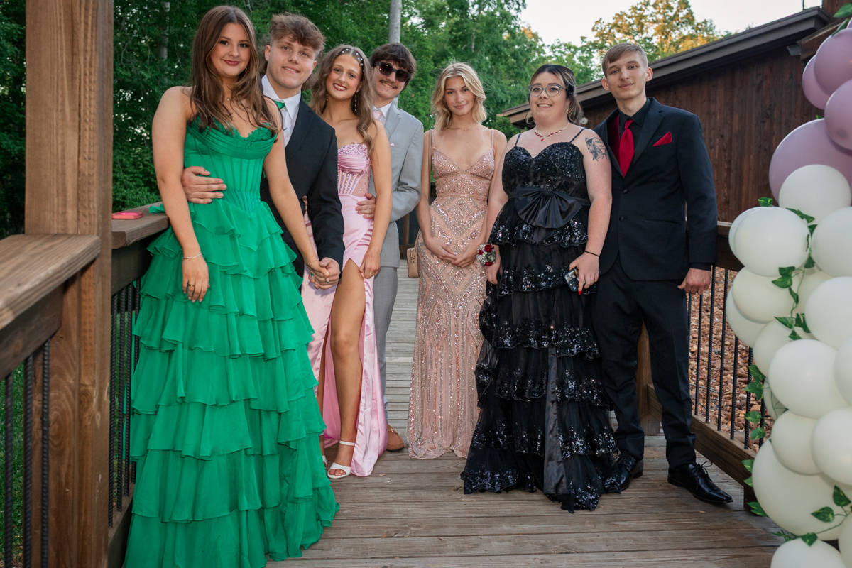 Group of seven teens in formal wear posing on a wooden deck outdoors, wearing colorful gowns and suits with balloons nearby.
