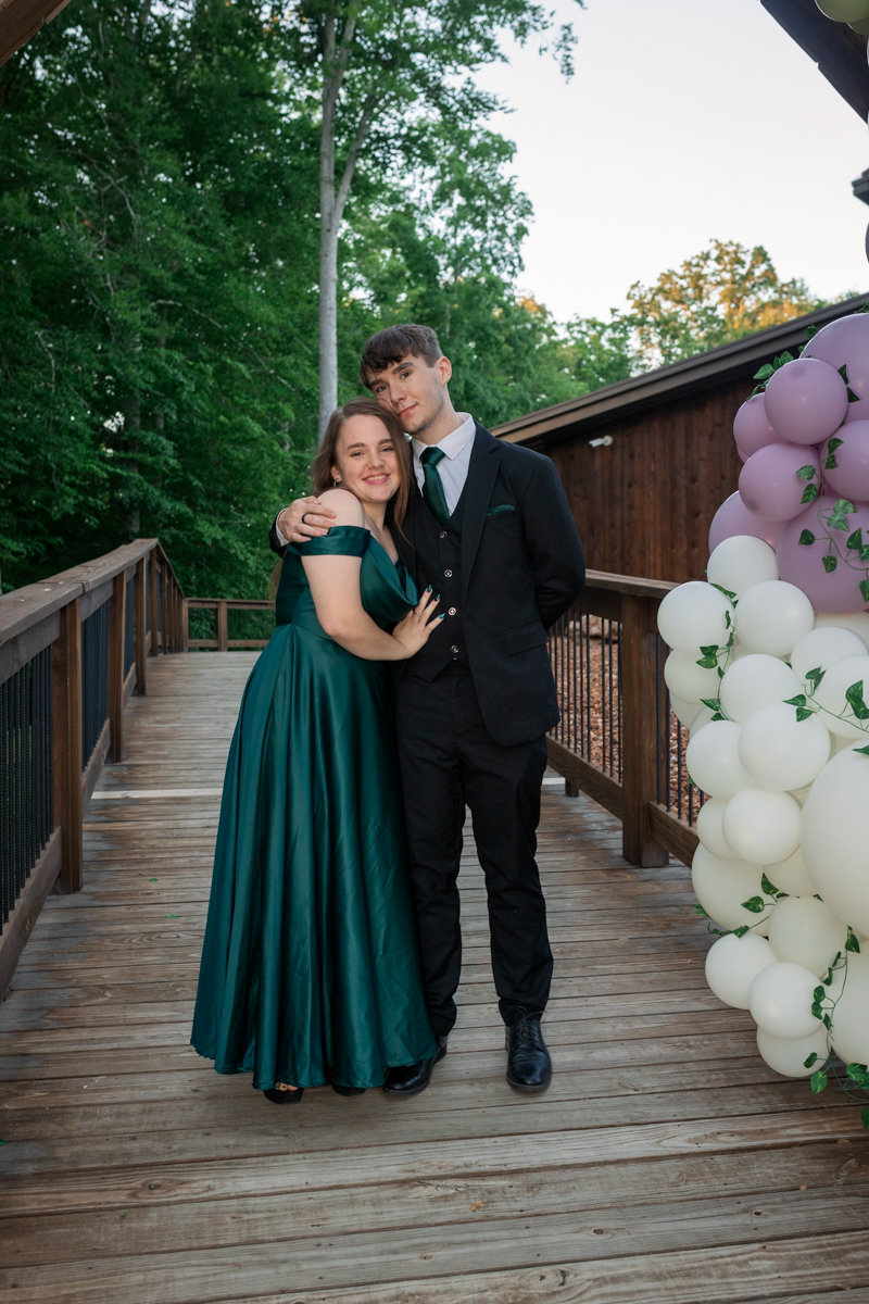 Couple posing on a wooden deck beside a pastel balloon arch; woman in a green gown and man in a black suit with a green tie, smiling.