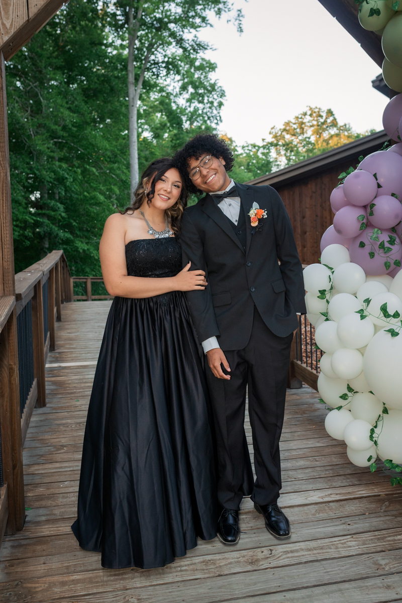Couple in formal wear posing arm in arm on a wooden deck with a purple and white balloon arch beside them.
