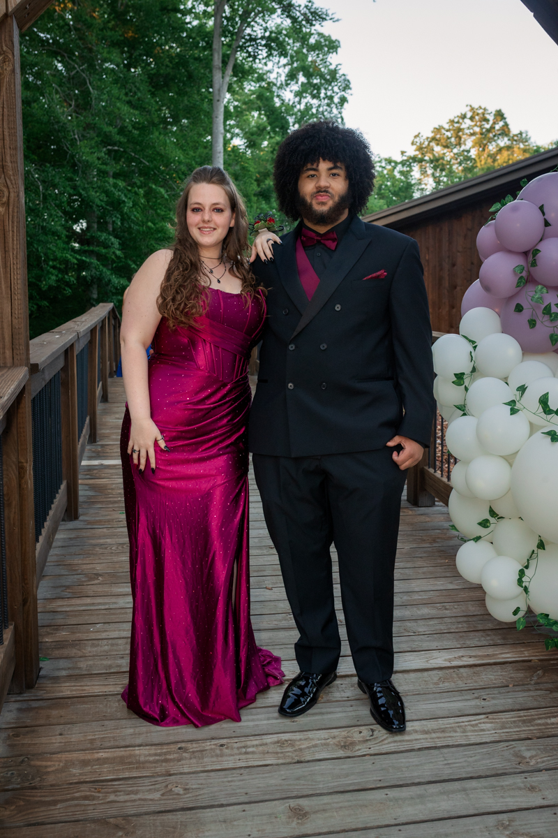 Couple in formal wear poses on a wooden bridge; woman in a magenta gown, man in a black suit with burgundy accents, balloons nearby.