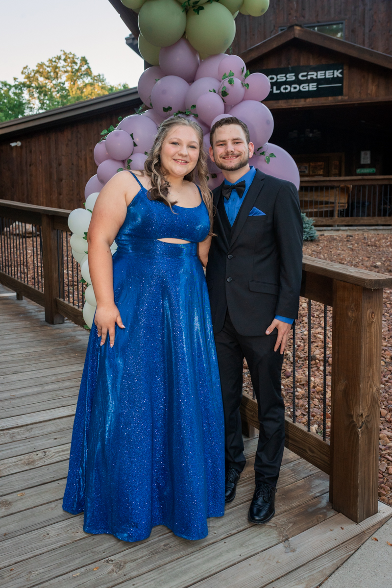 Young couple in formal attire posing beside a pink and green balloon arch outside a wooden lodge.
