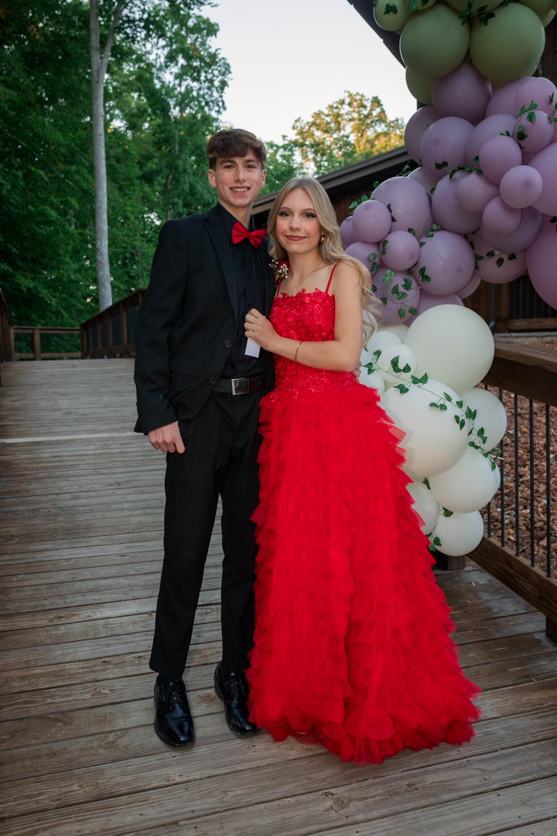 Young couple in formal wear posing on a wooden deck beside a balloon arch; woman in a red ruffled gown and man in a black suit with a red bow tie.