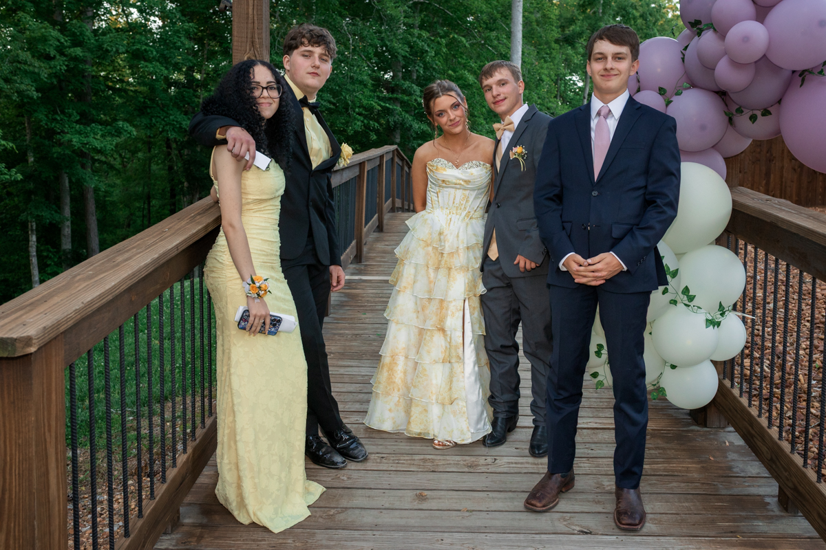 Group of five teenagers in formal wear posing on a wooden bridge with pink and white balloons in the background.
