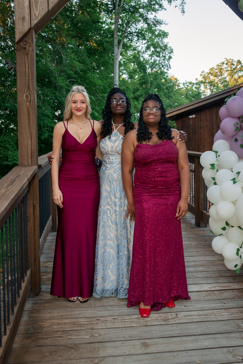 Three women posing in formal gowns on a wooden deck with balloons at an outdoor event; burgundy dress, light blue patterned dress, and magenta sequin dress.