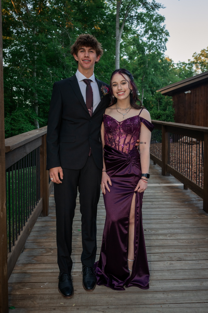 Two young adults in formal wear pose on a wooden boardwalk; man in a black suit with burgundy tie, woman in a purple embellished gown with a thigh-high slit.