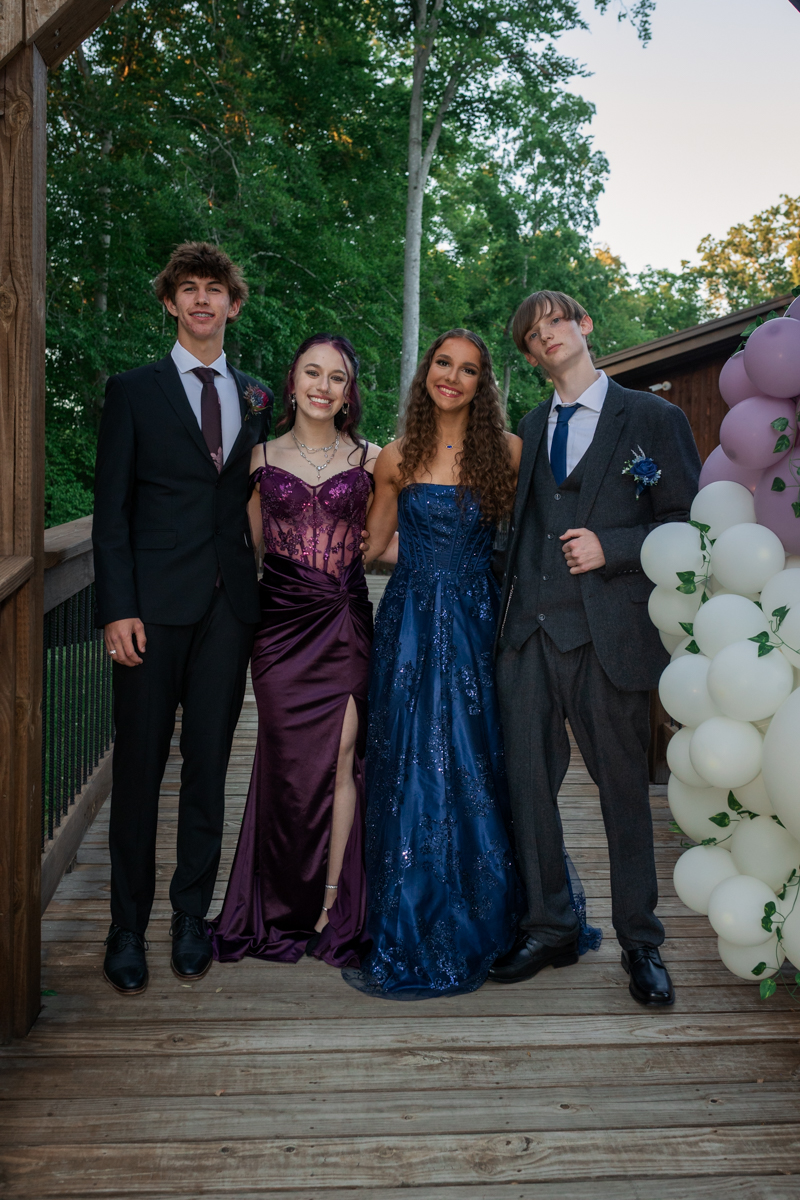 Four teens in formal wear posing on a wooden deck, with two girls in gowns (purple and blue) and two boys in suits, near a balloon arch outdoors.