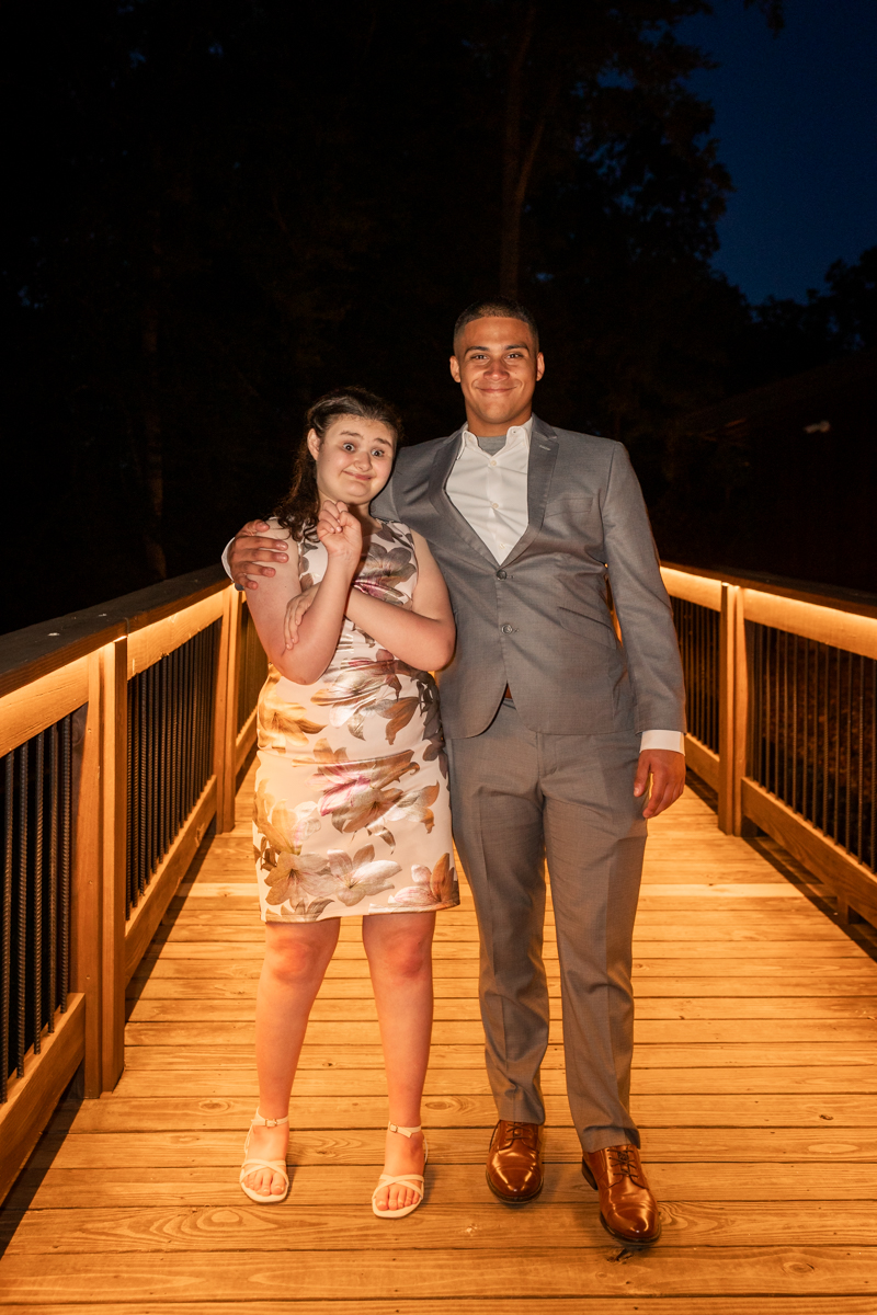 Two young adults pose arm-in-arm on a warmly lit wooden bridge at night; she wears a floral dress, he a gray suit.