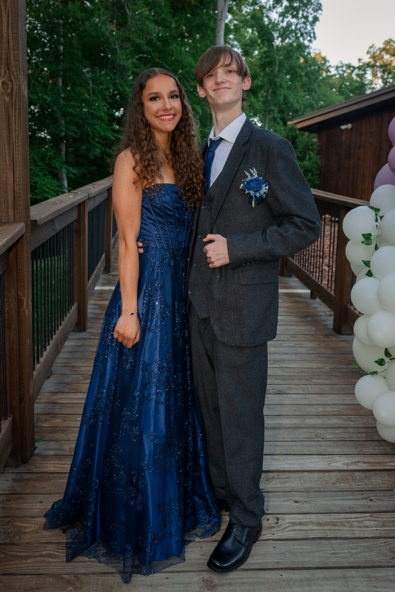 Smiling couple in formal wear posing on a wooden deck, girl in a navy blue gown and boy in a gray suit with a blue boutonniere, balloons nearby.