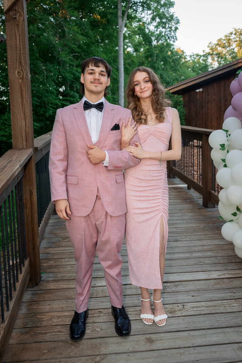 Couple dressed in pink formalwear posing arm-in-arm on a wooden deck with a balloon arch nearby at an outdoor event.