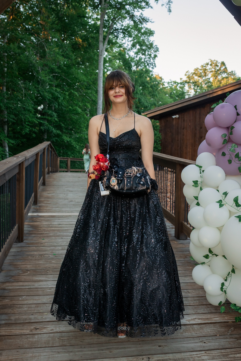 Young woman in a black sequined ball gown standing on a wooden bridge with balloons nearby.