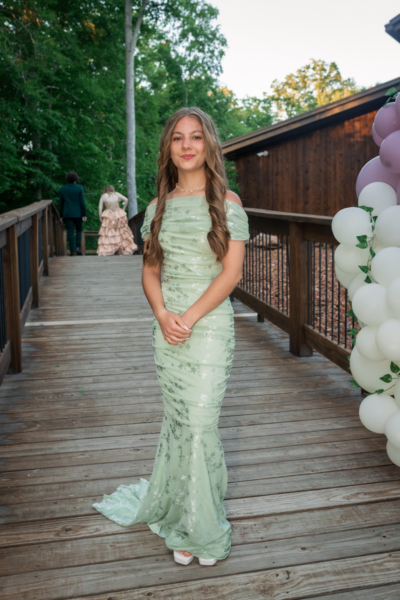 Teenage girl in a mint green off-the-shoulder formal dress standing on a wooden bridge, posing with hands clasped, pastel balloons on the right at an outdoor event.