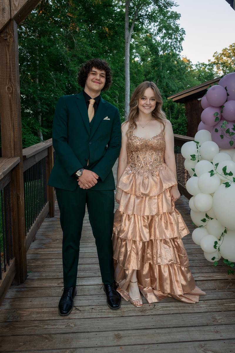 Two teens in formal attire stand on a wooden bridge with balloons beside them.