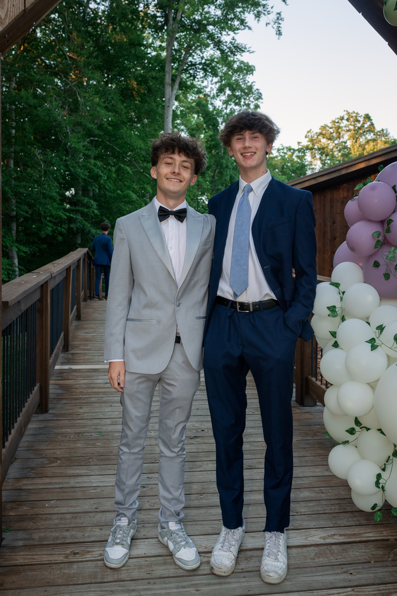 Two young men in suits stand on a wooden walkway outdoors, smiling, with pastel balloon arrangements nearby.