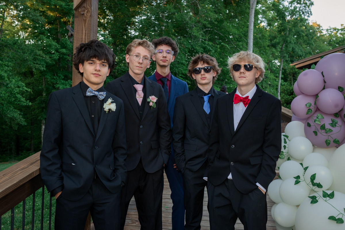 Group of five young men in suits posing on a wooden deck with pastel balloon decor, outdoors at a formal event like prom or a party.
