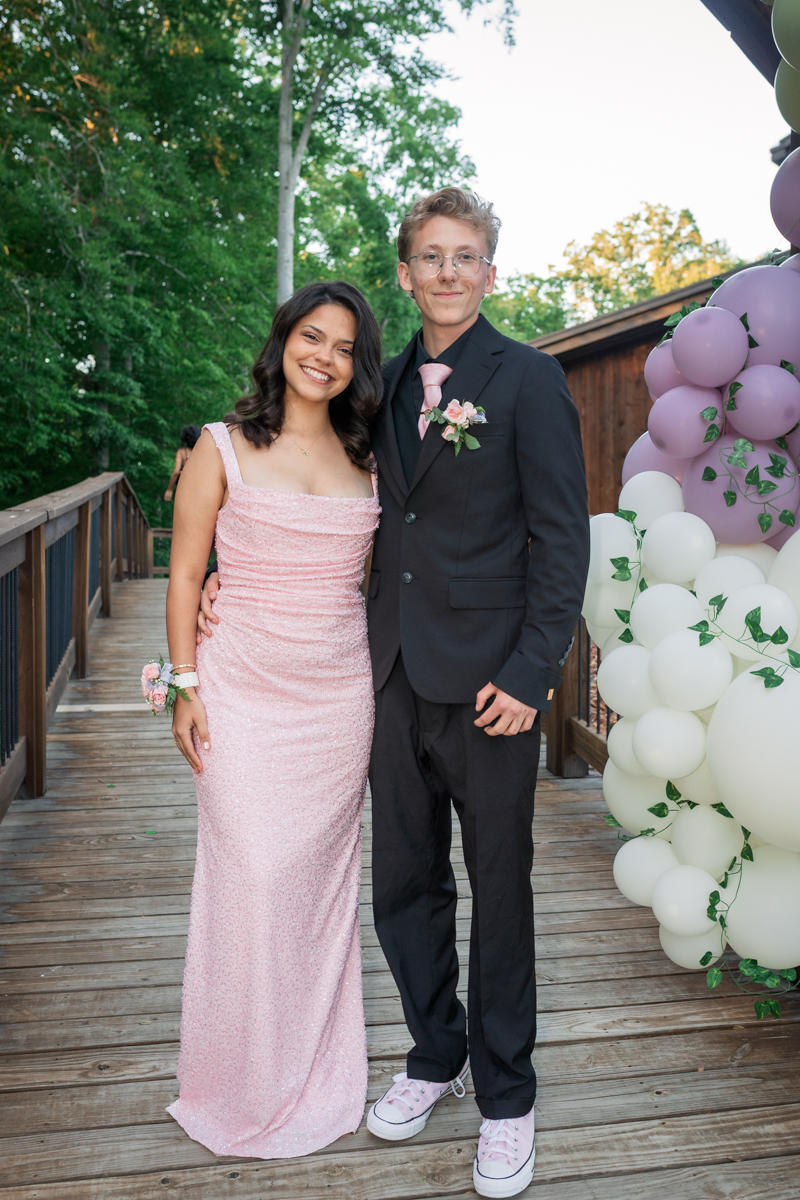 Couple posing on a wooden deck outdoors; woman in a pink sequin gown and corsage, man in a black suit with pink tie and boutonniere, pastel balloon arch in the background.