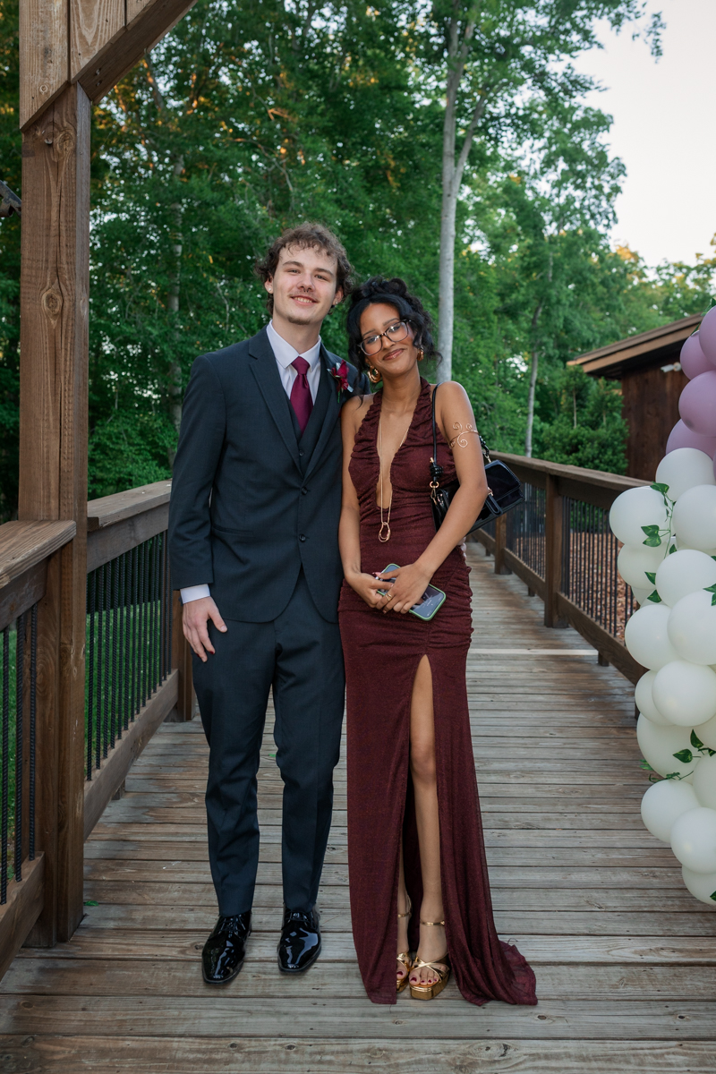 Young couple posing on a wooden outdoor deck in formal attire, a man in a dark suit and tie and a woman in a burgundy gown with a high slit, smiling at the camera.