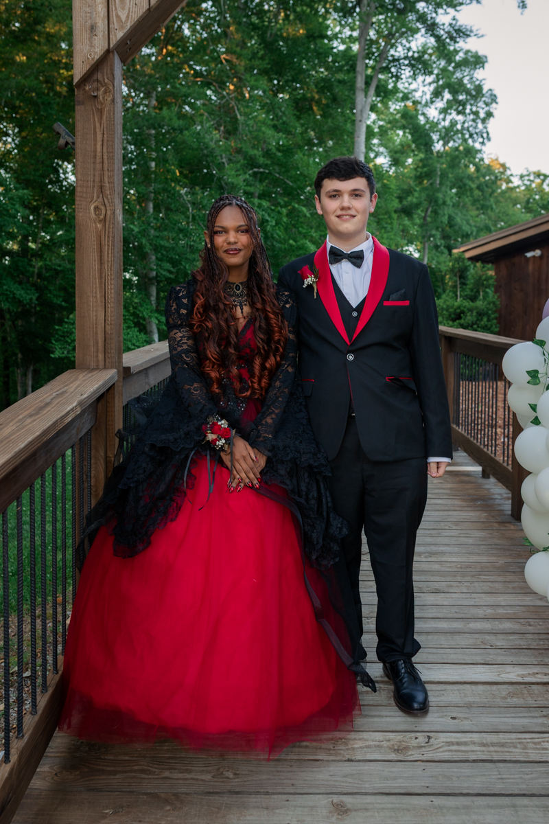 Couple in formal attire posing on a wooden deck outdoors; woman wears a red and black ball gown with lace cardigan, man in a black suit with red accents and bow tie.