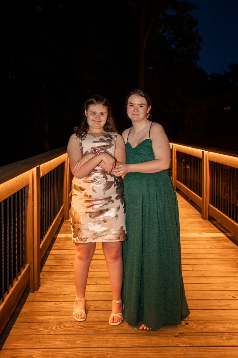 Two young women pose together on a lit wooden deck at night, one in a floral dress and the other in a green gown, holding hands.