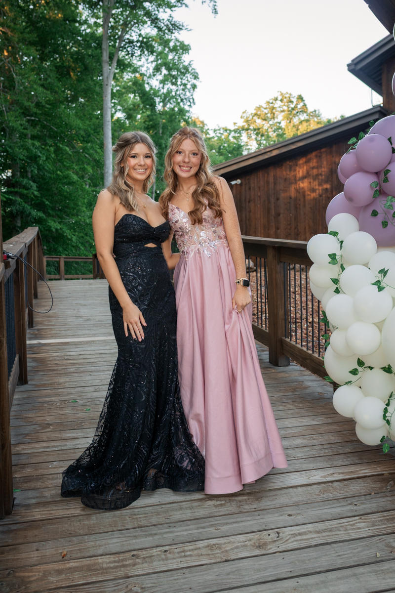 Two women in formal gowns posing on a wooden deck with lavender and white balloons beside them, outdoors at a celebration.