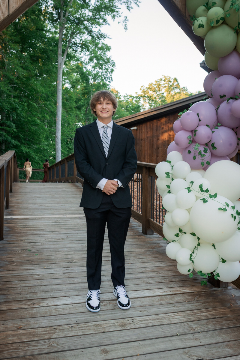 Young man in a black suit and striped tie stands on a wooden walkway beside a pastel balloon arch outdoors.