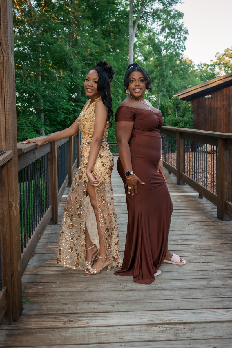Two women in formal gowns pose on a wooden outdoor deck with green trees in the background.