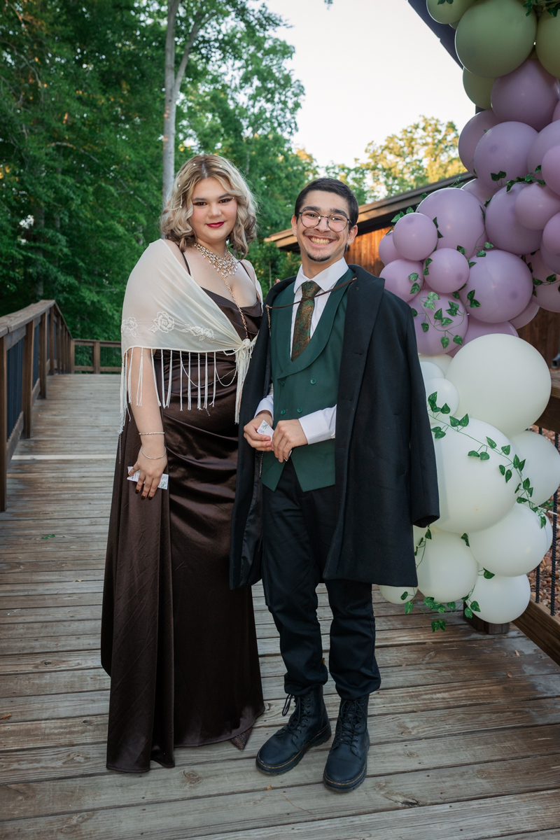 A couple in formal attire stands on a wooden deck beside a lavender and white balloon arch at an outdoor event, smiling at the camera.