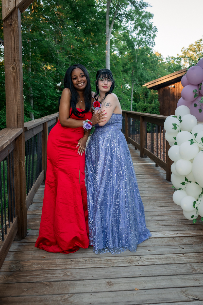 Two women in formal gowns stand on a wooden deck, smiling, one in a red dress and the other in a lavender sequined gown with balloons nearby.