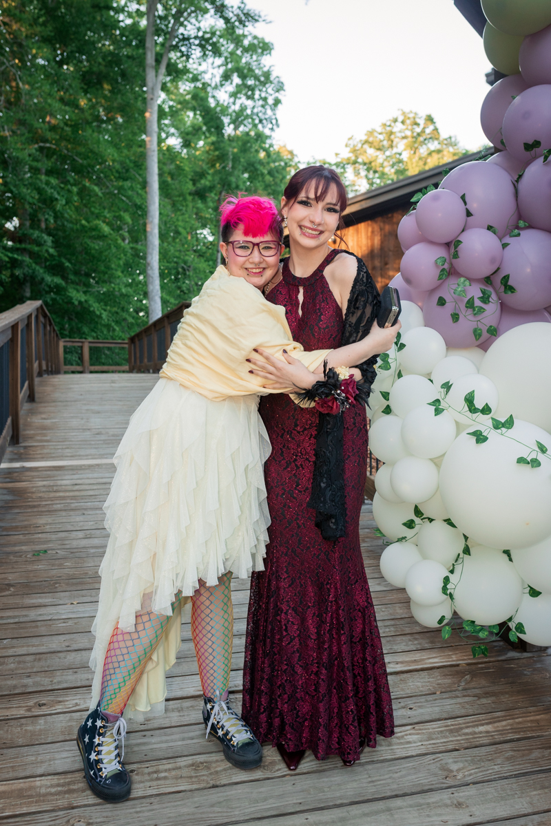 Two friends hug and smile on a wooden deck beside a pastel balloon arch at an outdoor celebration.