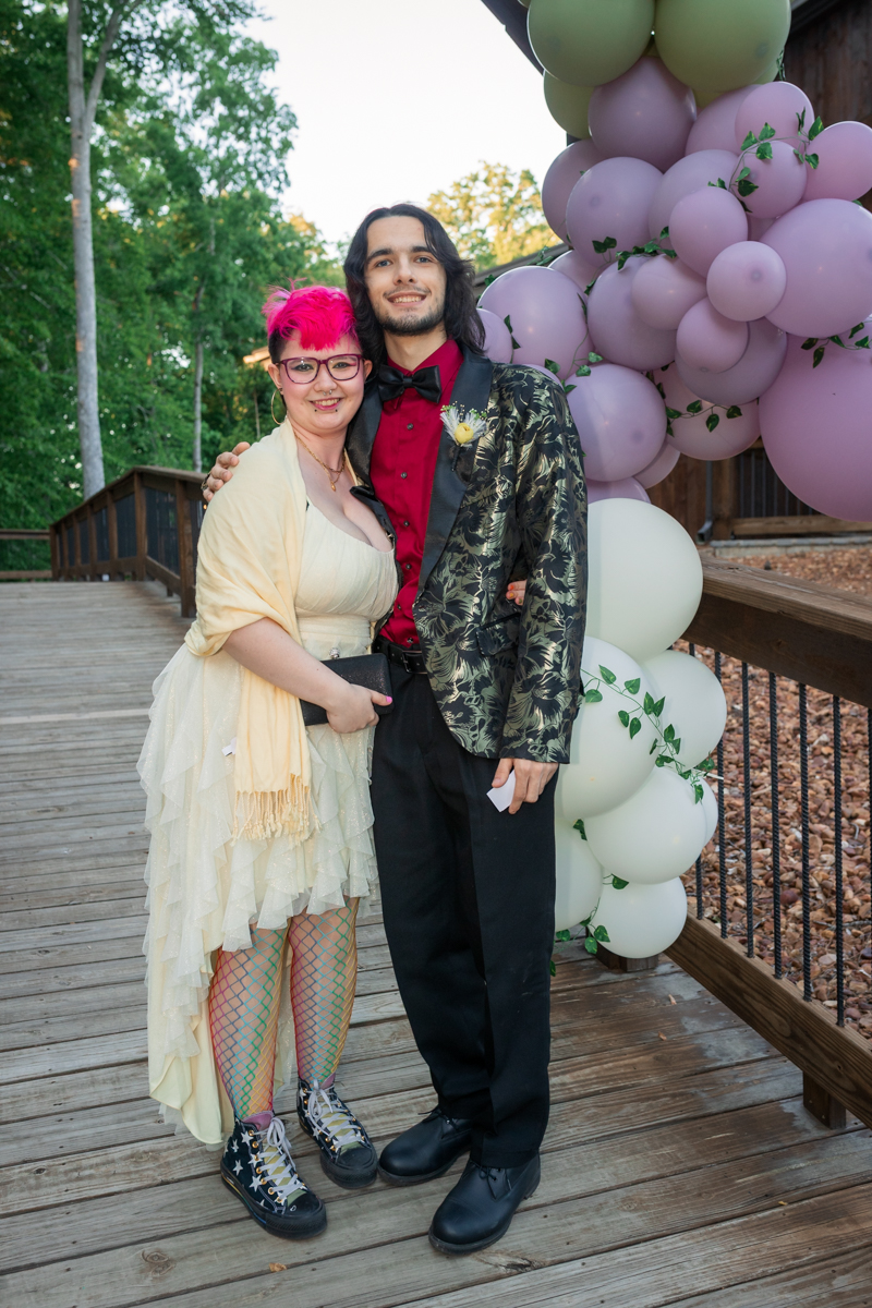 Couple posing together at a celebration on a wooden deck with a pastel balloon arch nearby.
