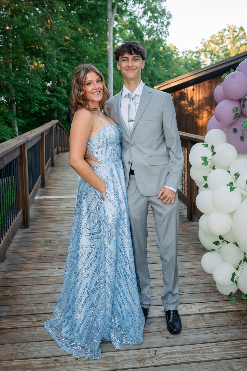 Young couple posing outdoors on a wooden deck; girl wears a blue sequined gown and boy a light gray suit, with balloons nearby.