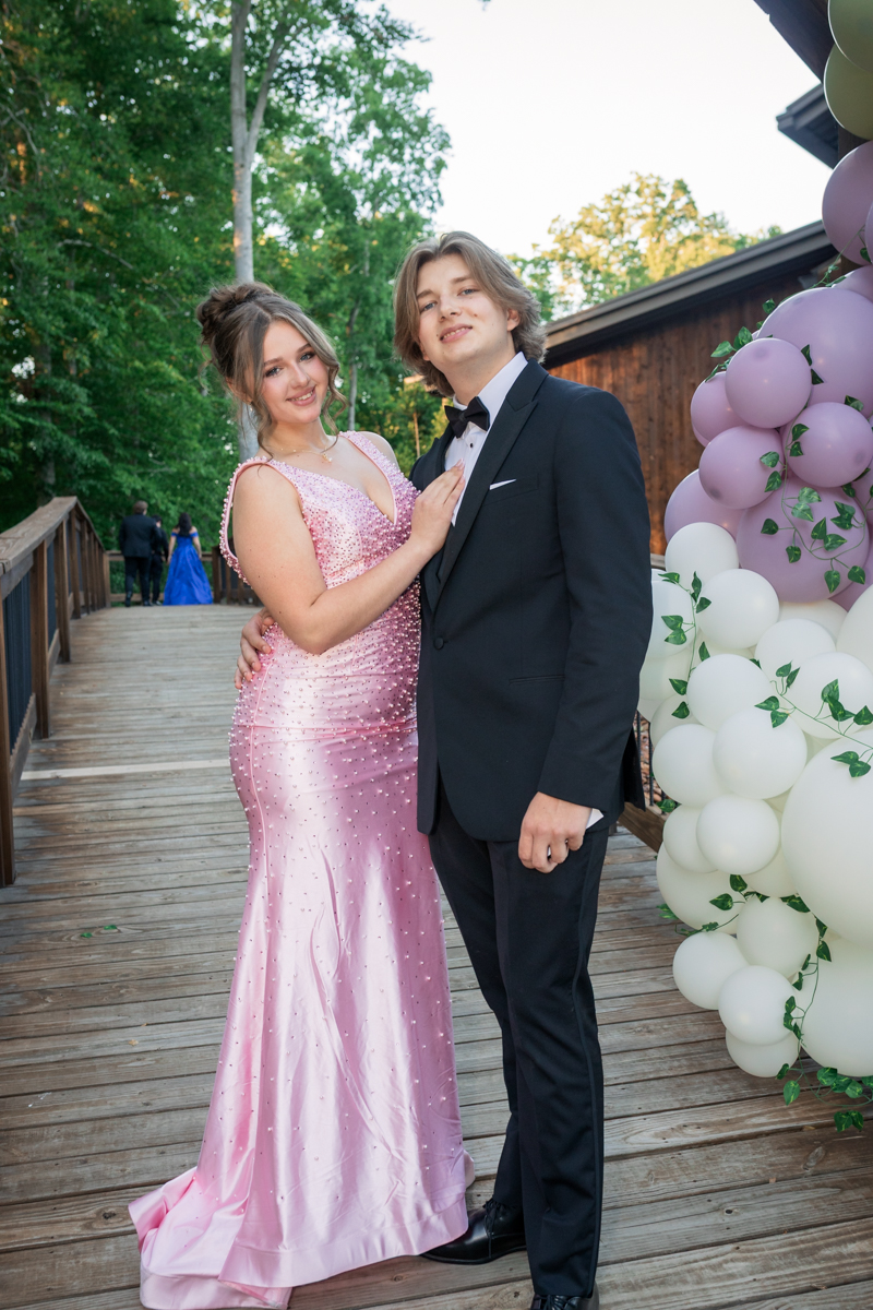 Couple dressed formally posing on a wooden deck with pink dress and black tuxedo beside a balloon arch at an outdoor event.