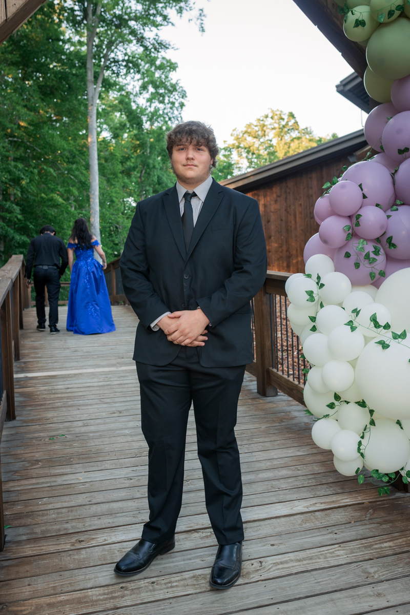 Young man in a black suit and tie stands on a wooden deck beside a pastel balloon arch at an outdoor event.