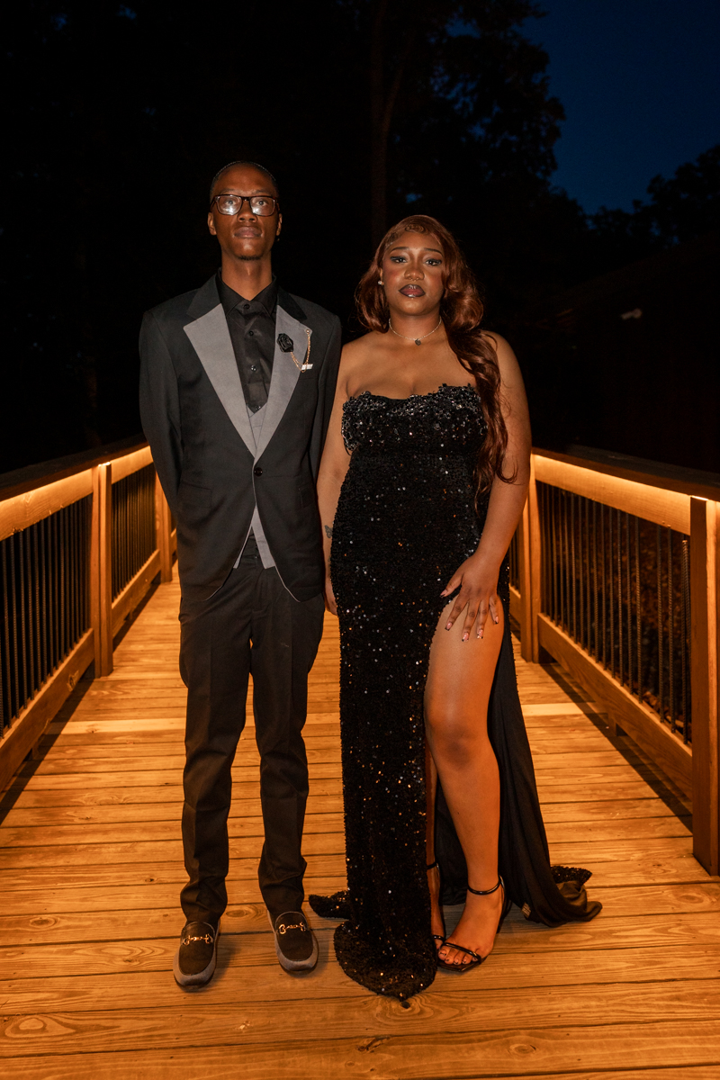 Two formally dressed individuals pose on a wooden bridge at night, the woman in a black sequined gown with a high slit and the man in a black suit with gray lapels.